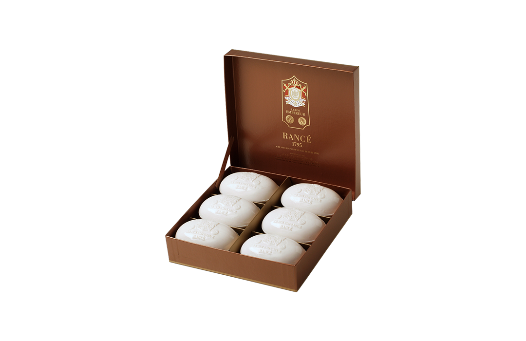 An open luxurious brown box containing six Rance Le Roi Empereur Boxed Soap, each embossed with a decorative design, set against a striped background.