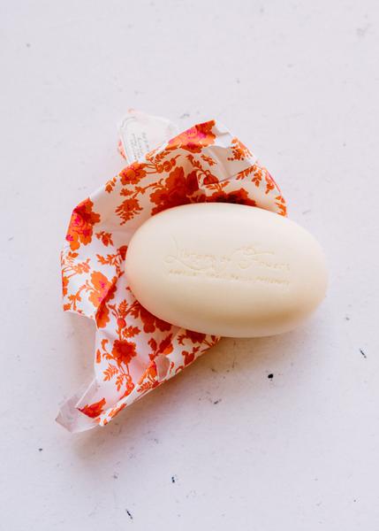 A bar of Library of Flowers FIELD & FLOWERS PERFUMED SOAP from Margot Elena, with floral embossment, partially wrapped in paper with a red and orange floral design, placed on a white textured background. The soap is enriched with Shea Butter for deep moisturization.