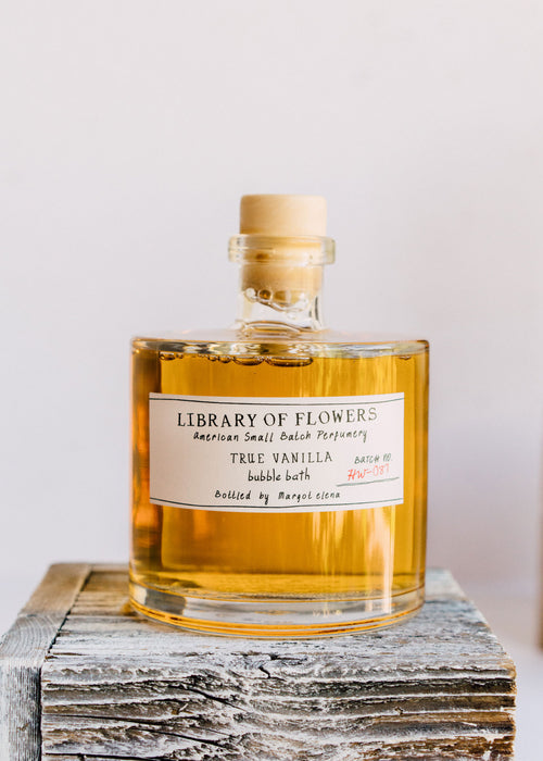 A clear glass bottle of Margot Elena's Library of Flowers True Vanilla Bubble Bath atop a rustic wooden block, positioning the well-lit label front and center against a neutral background.