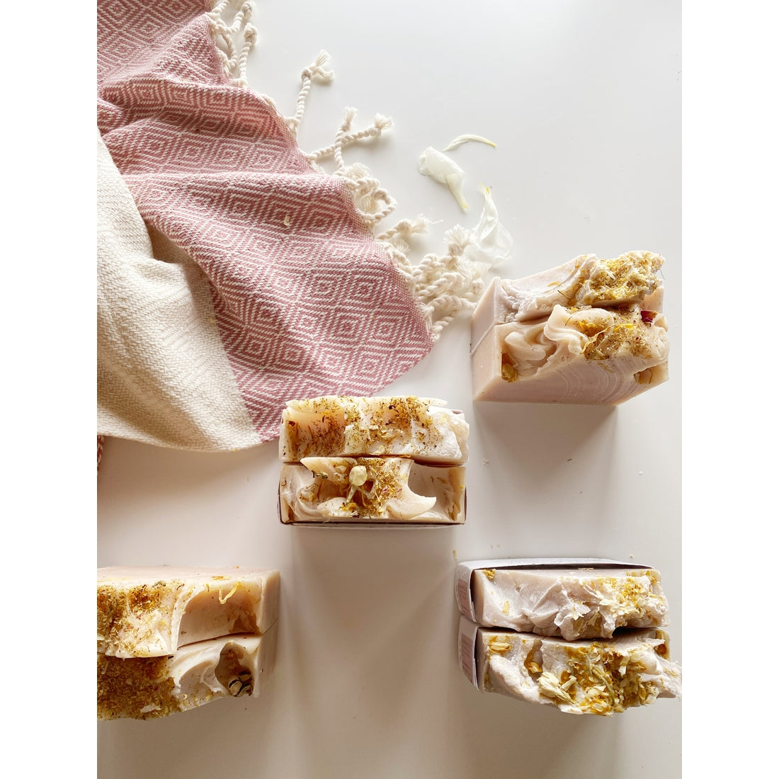 Handmade Lilac Soap Bars with flower petals and gold flakes, arranged neatly beside a pink patterned towel on a white surface, with some creamy sustainable soap mixture artistically splattered around.