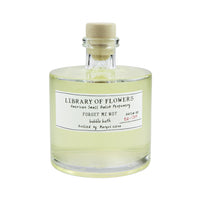 A clear glass bottle of Margot Elena's Library of Flowers Forget Me Not Bubble Bath with a cork stopper. The label reads "forget me not" and indicates it's from American small batch perfumery, featuring silky