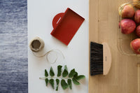 Flat lay of a red notebook, twine, an Andrée Jardin Mr. & Mrs. Clynk Natural Table Brush made from 100% natural fibers, and a sprig of green leaves on a white surface next to a glass box filled with red apples