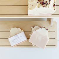 Handmade soaps displayed on wooden shelves, featuring labels describing the ingredients. One soap, named "SOAK Bath Co. - Rosé All Day Soap Bar," is floral-topped, enhancing its handmade, sustainable allure.