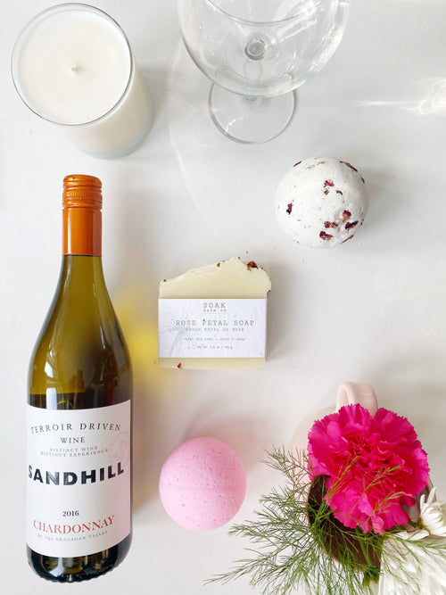 A flat lay featuring a bottle of Sandhill Chardonnay, a rose petal bath bomb, SOAK Bath Co. - Rose Petal Soap, a scoop of ice cream, a candle, a wine glass.