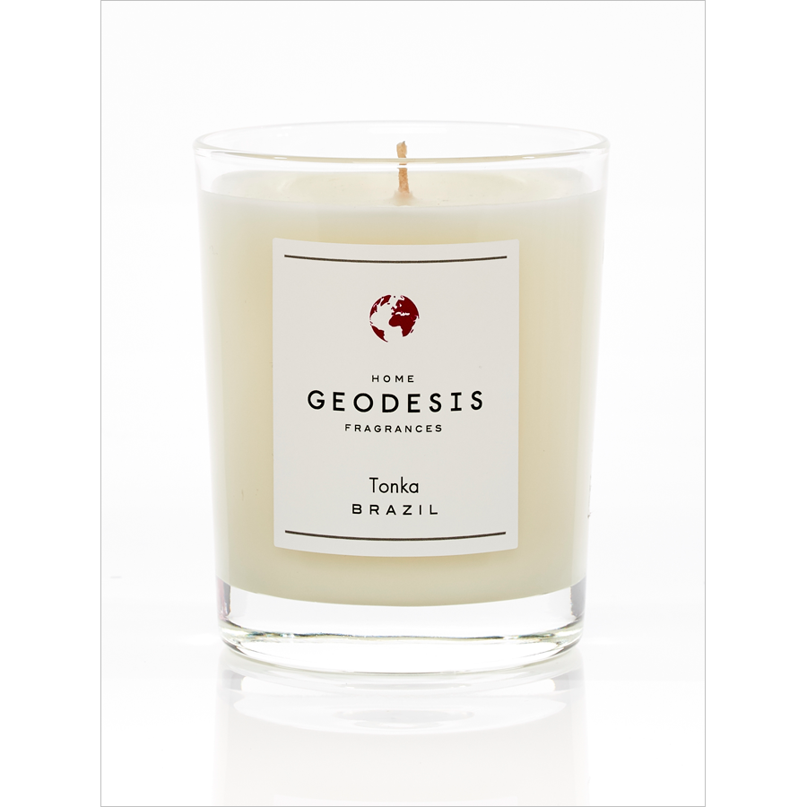 A large white candle in a clear glass container labeled "Geodesis" with "Tonka Brazil" and a burning time of 50 hours written underneath, featuring a small red and white.