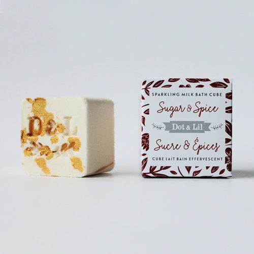 A Dot & Lil Sugar + Spice Sparkling Milk Bath Cube with gold flakes and the letter "d" imprinted, next to its packaging labeled "Dot & Lil." The background is plain white.