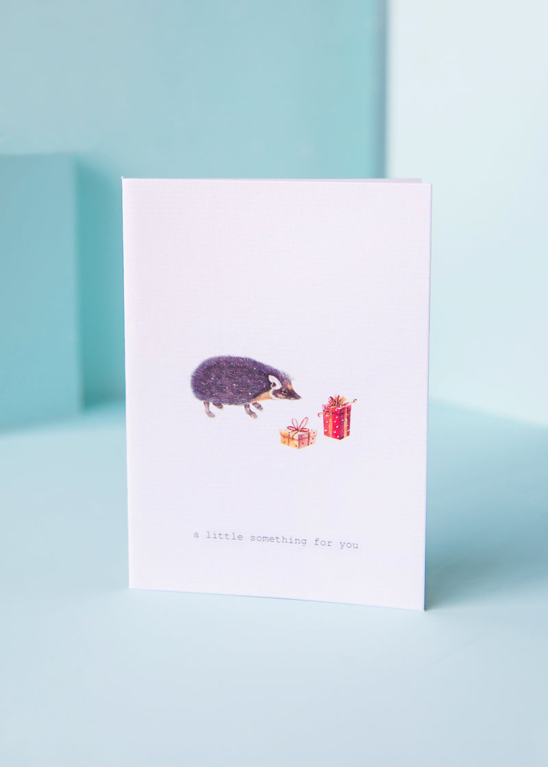 A TokyoMilk Little Something Greeting Card on a light blue textured paper background featuring a printed image of a hedgehog and two gift boxes with the text "a little something for you" beneath. Made by Margot Elena.