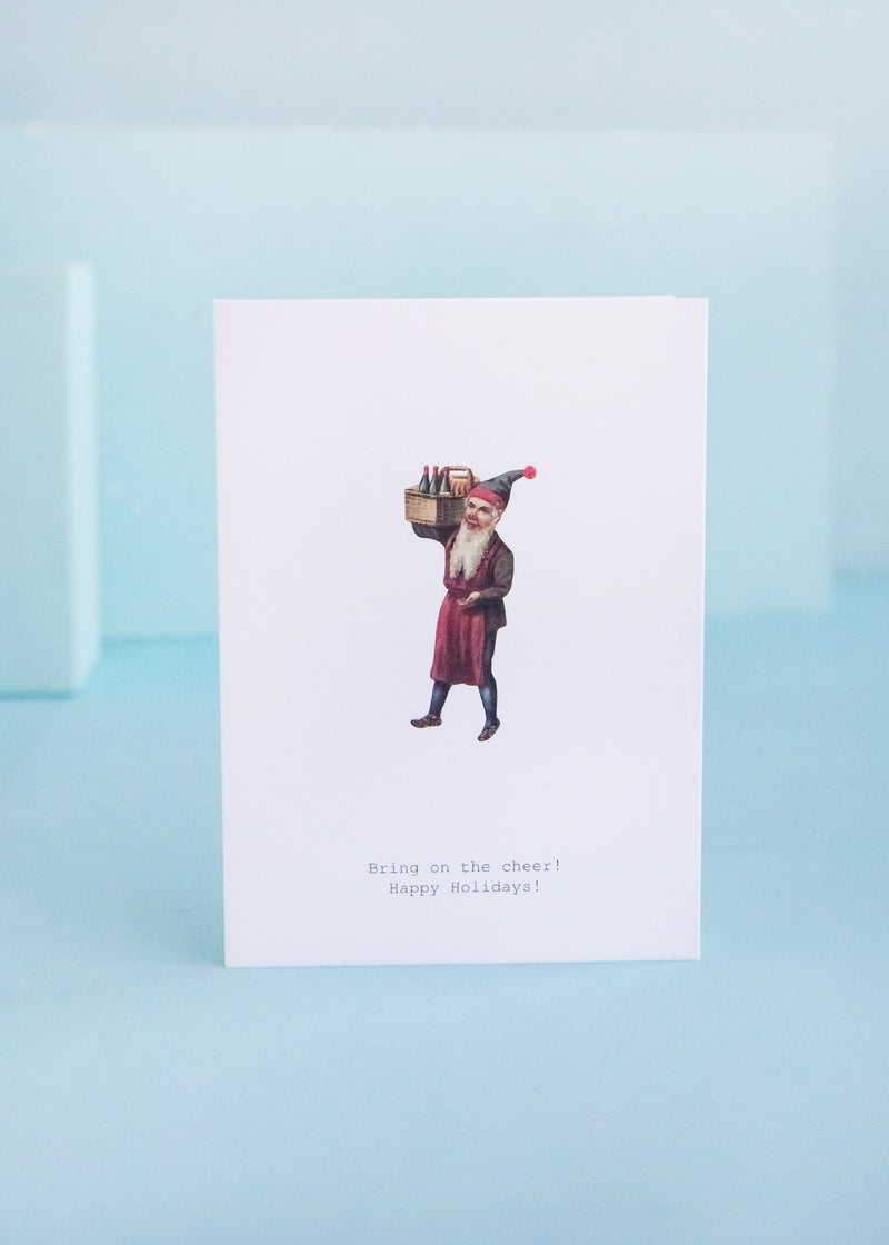 A Margot Elena TokyoMilk Greeting Card featuring a vintage illustration of a man carrying a stack of gifts on his head, with the text "bring on the cheer! happy holidays!" on a textured paper background.