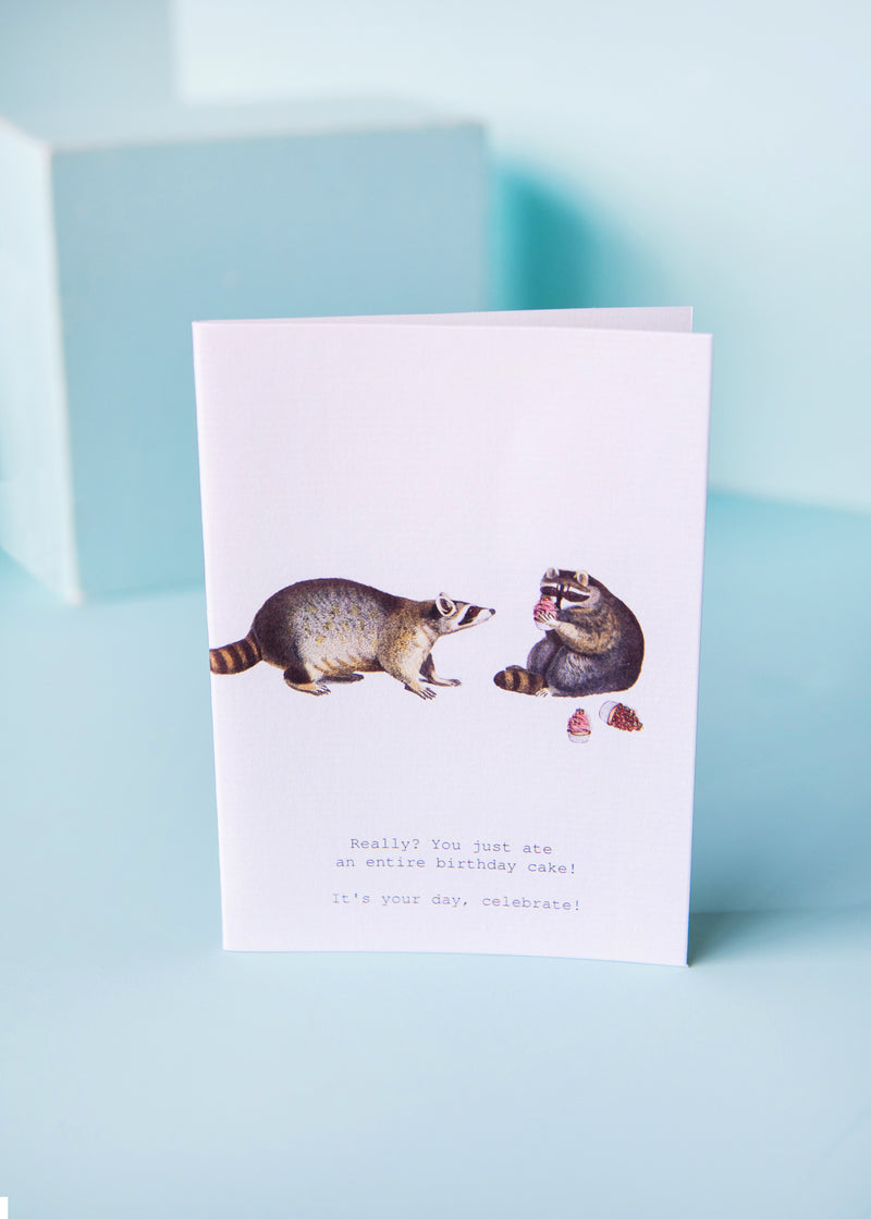A TokyoMilk Greeting Card - Raccoons Your Day Greeting Card features two illustrated raccoons, one looking surprised at a bite taken from a slice of cake, with the caption "Really? You just ate an entire birthday cake! It’s your." Created by Margot Elena.