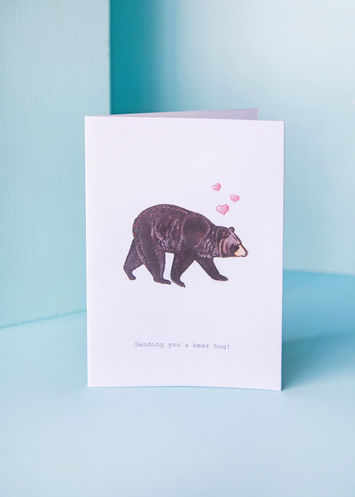 A TokyoMilk Bear Hug Greeting Card crafted from textured paper stands against a soft blue background. It features an illustration of a brown bear with three pink hearts above its head and the caption "Sending you a bear hug!" by Margot Elena.