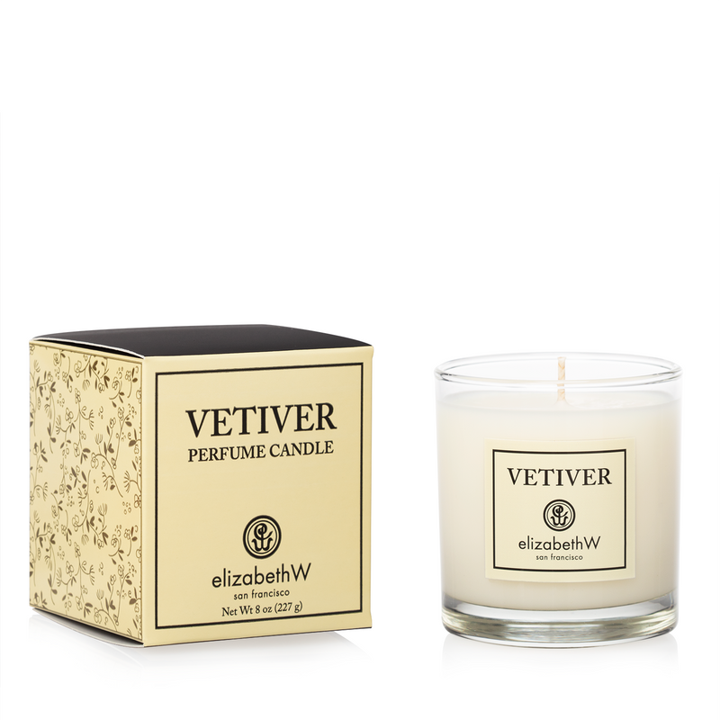 A elizabeth W Signature Vetiver Candle by Elizabeth W in a clear glass container with a lit wick, next to its decorative yellow and black packaging box labeled with the product name.