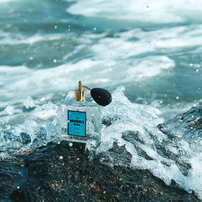 A perfume bottle labeled "Musgo Real" sits on a wet, rocky surface. Waves crash around it, sending splashes of water into the air. The background features a blurred view of the ocean.