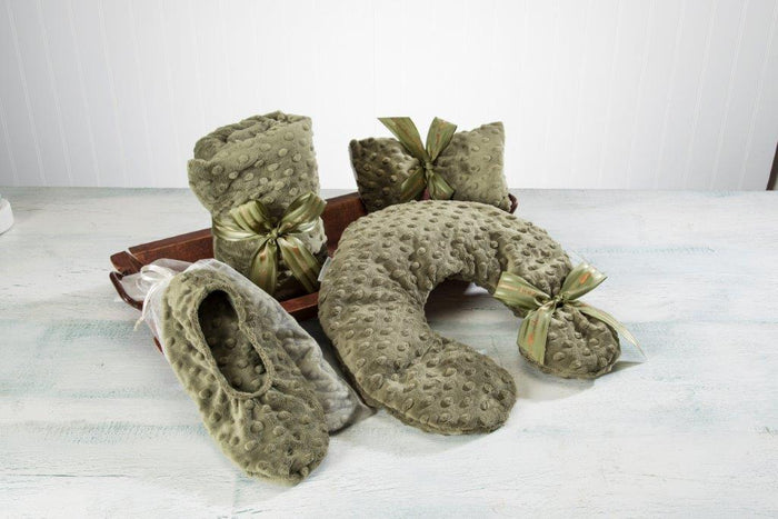 A set of cozy green textured products displayed on a light surface, including slippers, a neck pillow, and two rolled blankets tied with ribbons, on a wooden tray.