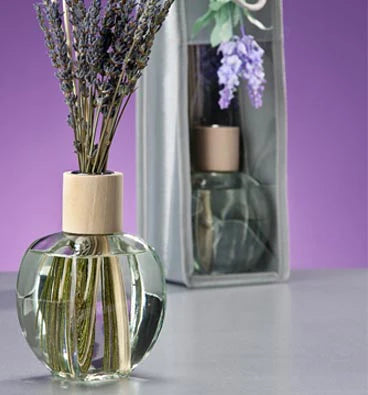 A small round glass diffuser with lavender stalks is in the foreground. Behind it is a boxed diffuser with a similar design. The background is a gradient of purple shades.