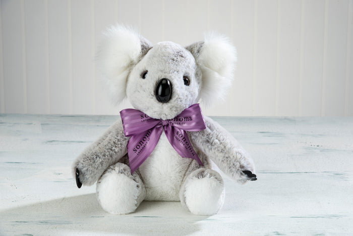 A plush koala with white fluffy ears and a purple ribbon sits on a light wooden surface against a white paneled background.