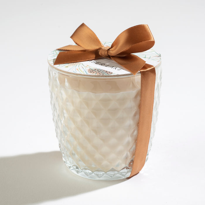 A textured glass jar candle with a creamy white wax inside and a gold ribbon tied in a bow around the lid. The label on the lid is partially visible. The background is plain white.