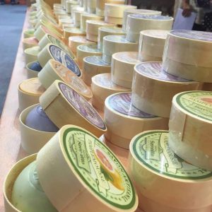 Rows of rounded boxes of handmade soap are arranged diagonally on a wooden surface. Each box has a colorful label, indicating different scents and types of soap. The background is out of focus.