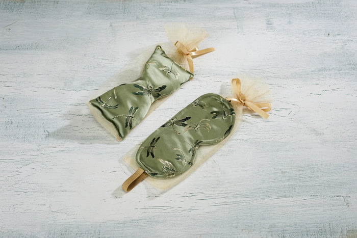 Two green and gold embroidered eye masks with ribbon ties are neatly placed on a light wooden surface. Each mask is adorned with a delicate dragonfly design.