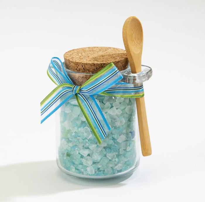 A glass jar filled with blue bath salts, topped with a cork lid. A striped blue and green ribbon is tied around the jar. A small wooden spoon is attached to the side. The background is plain white.