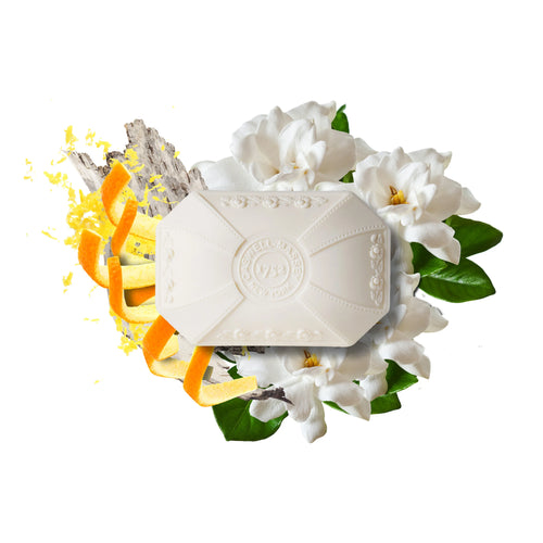 Caswell-Massey Gardenia Bar Soap features floral designs and embossed text, surrounded by gardenia flowers, green leaves, orange peel, zest, and driftwood. This triple-milled, plant-based glycerin soap is shown on a white background.