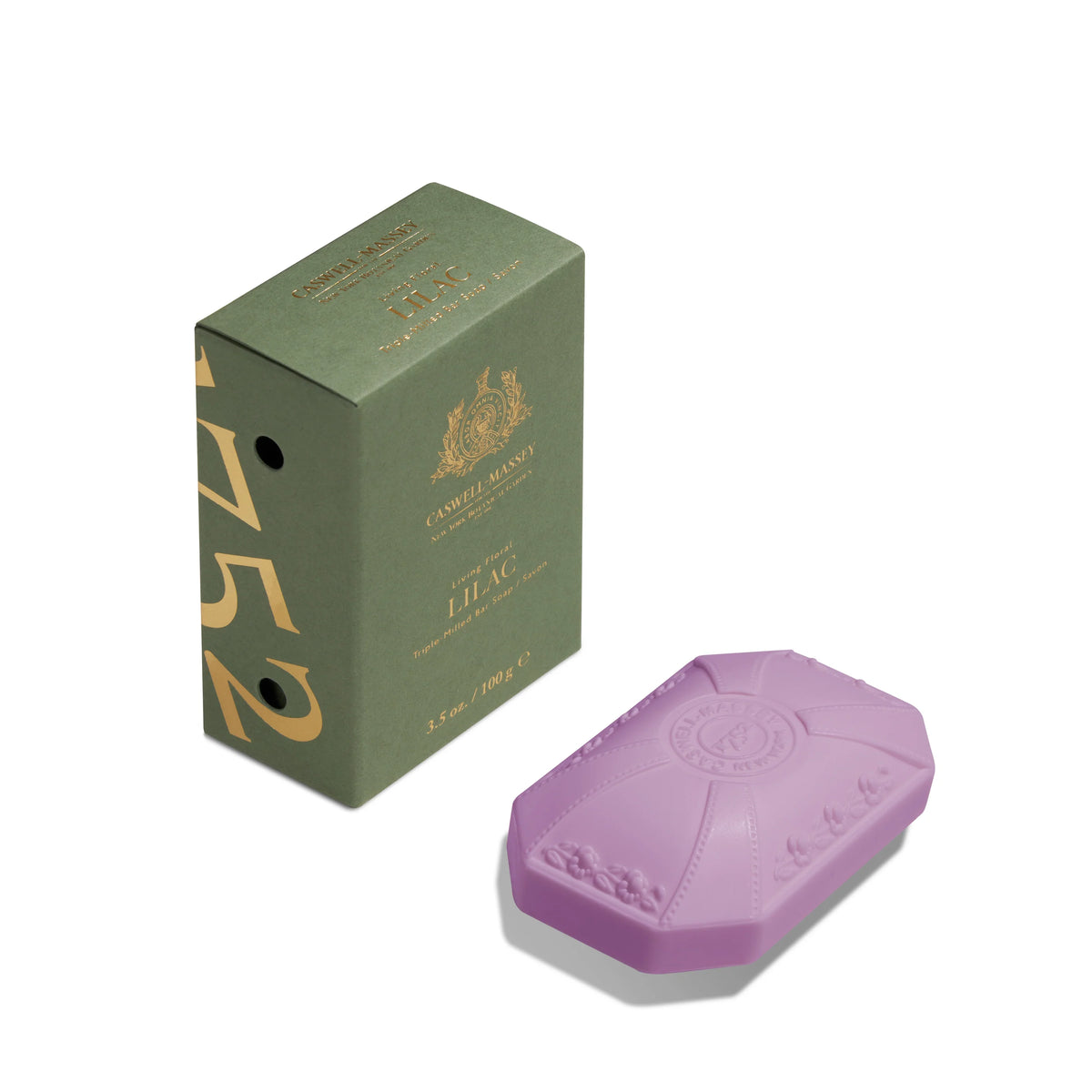 A lavender-colored bar of Caswell - Massey Lilac Luxury Bar Soap lying in front of its green packaging box, which features elegant gold text and design elements. The soap is embossed with ornate details and a