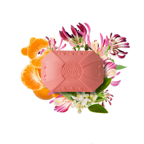 The Caswell-Massey Honeysuckle Bar Soap, a pink triple-milled soap, is displayed in front of vibrant flowers and orange slices on a white background for a fresh, colorful look.