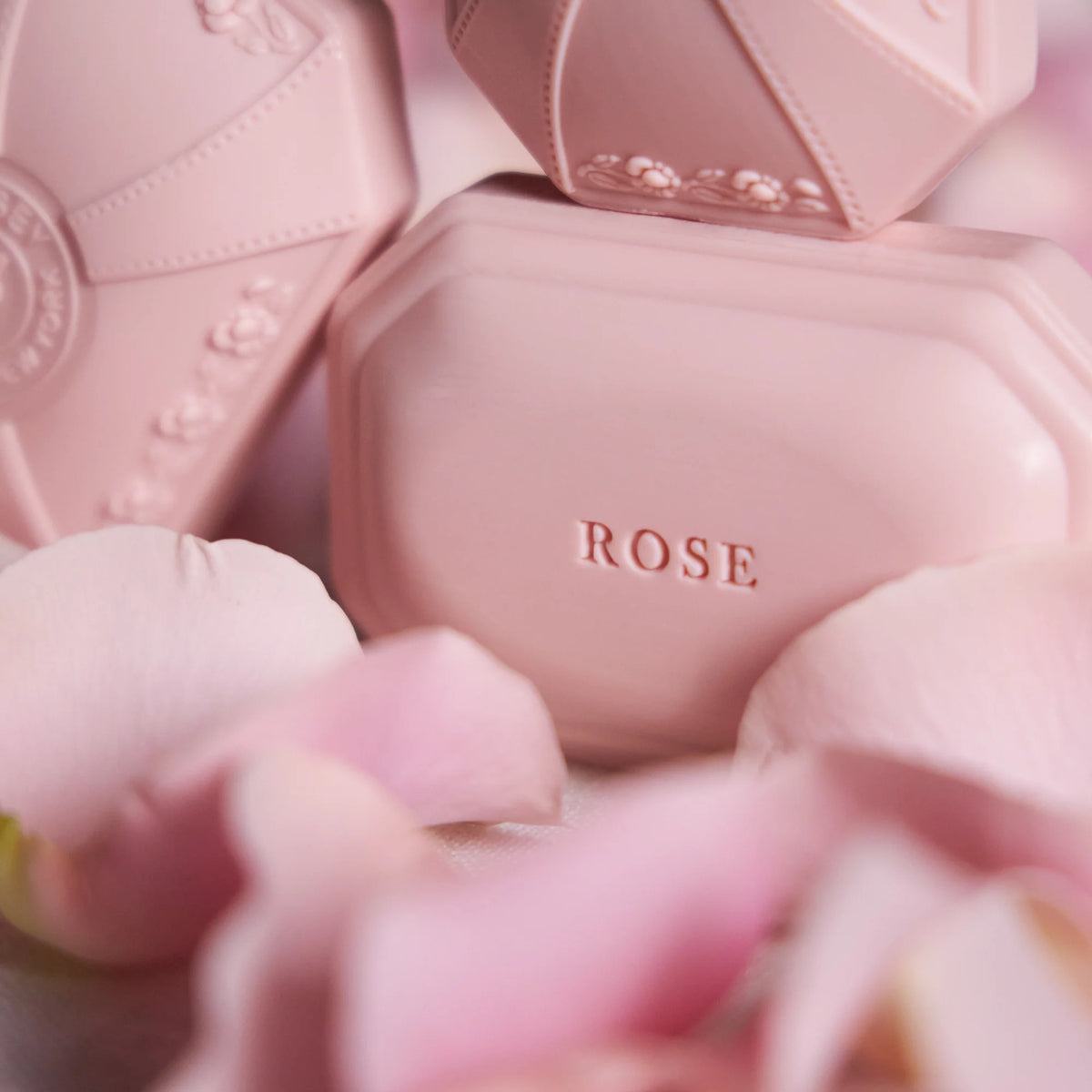 Close-up of soft pink skincare products with embossed details, surrounded by delicate rose petals. The word "rose" is prominently displayed on a Caswell Massey Rose Luxury Bar Soap 100g.