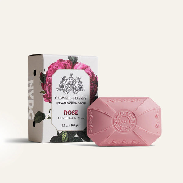The Caswell-Massey Rose Luxury Bar Soap 100g sits next to its floral box with a rose illustration and the brand's logo. The plant-based glycerin soap is embossed with the Caswell-Massey seal.