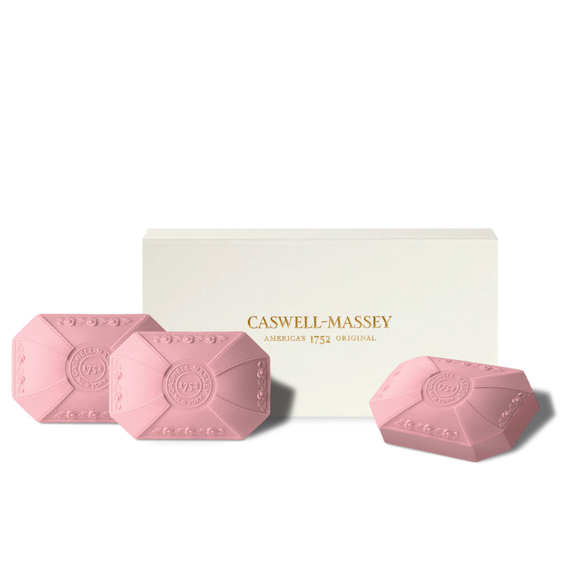 Three pink, hexagonal, triple-milled soaps with embossed designs beside a white box labeled "Caswell Massey Rose Three-Soap Set America's 1752 Original" on a light background.