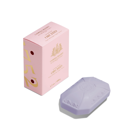 A Caswell-Massey Orchid Bar Soap, a lavender triple-milled bar with an embossed design, sits next to its pink "Orchid" box on a white background.