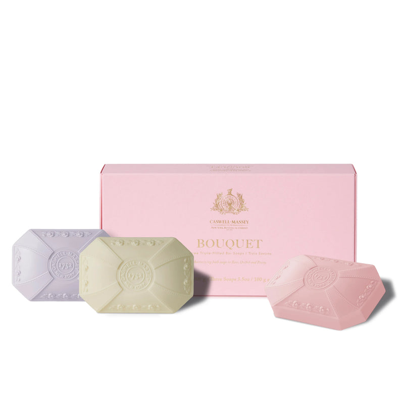 Three floral scented, luxurious soaps in pastel shades of pink, green, and purple, with embossed designs, displayed in front of a light pink rectangular box labeled "Caswell - Massey Three-Soap Set - Bouquet.