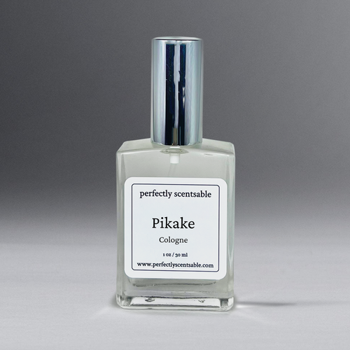 A clear glass bottle of cologne with a silver cap is centered in the image. The bottle has a white label that reads "Perfectly Scentsable Pikake Perfume Oil - 1oz Cologne Spray," featuring a jasmine blend inspired by the Pikake flower, with a website URL at the bottom. The background is a gradient of gray.