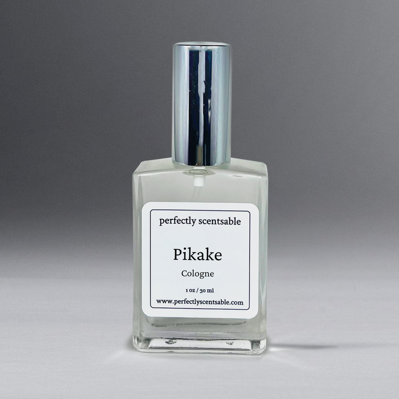 A clear glass bottle of cologne with a silver cap is centered in the image. The bottle has a white label that reads "Perfectly Scentsable Pikake Perfume Oil - 1oz Cologne Spray," featuring a jasmine blend inspired by the Pikake flower, with a website URL at the bottom. The background is a gradient of gray.