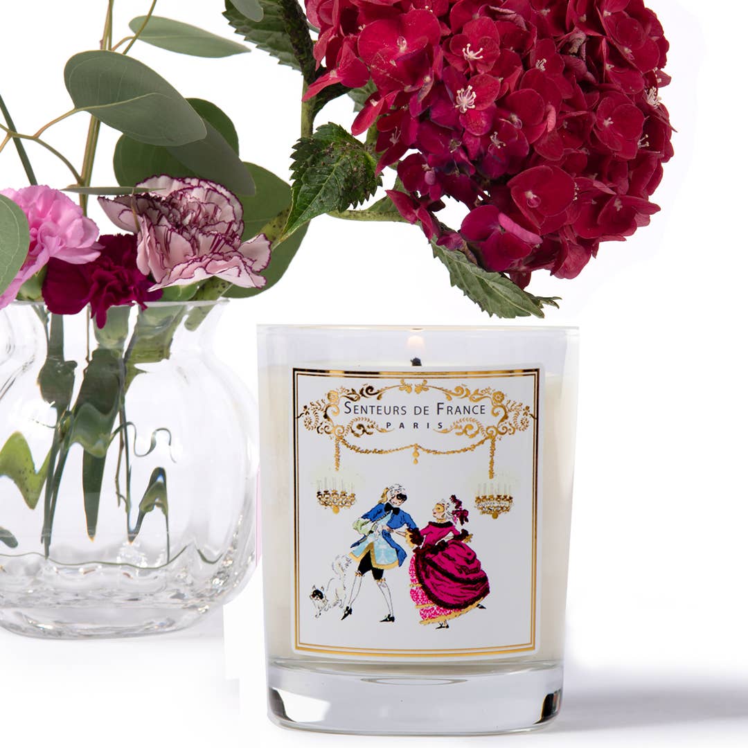 The Senteurs De France Violet Candle Royal Masquerade Ball, a white vegetable wax candle in a glass holder with a vintage label, sits beside a clear vase of red and pink hydrangeas and carnations.