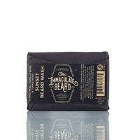 A black SUNRISE Beard Wash Bar from The Immaculate Beard showcases citrus, teak, vanilla, and musk notes, beard care benefits, ingredients list, and barcode—all reflected on a glossy white surface.
