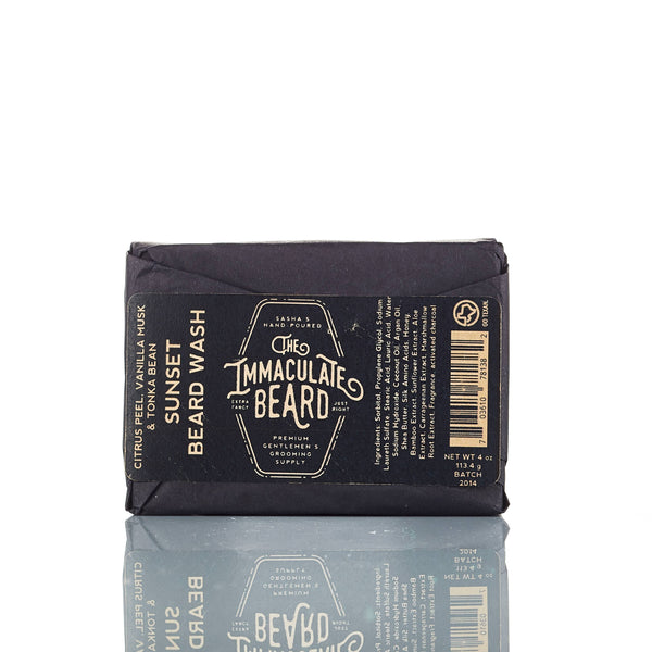 A black beard wash bar from The Immaculate Beard, wrapped in dark paper labeled "The Immaculate Beard SUNSET Beard Wash Bar," featuring scent, ingredient, and net weight info. The activated charcoal solid shampoo bar is reflected on a white surface.