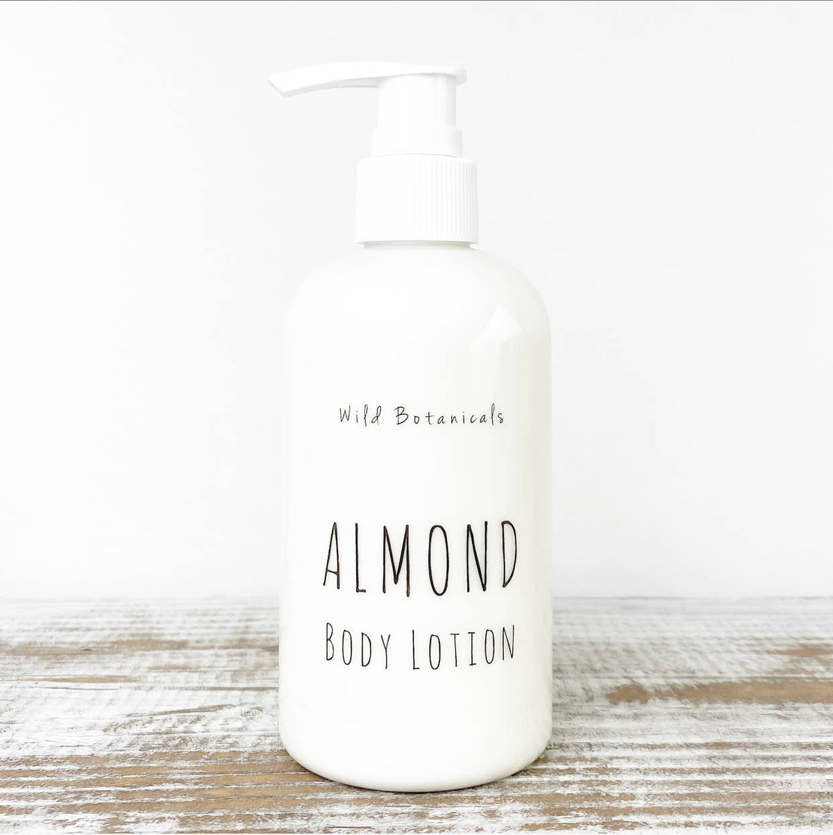 A white pump bottle labeled "Wild Botanicals Almond Lotion" stands on a rustic wooden surface with a neutral background. The label features simple, minimalist typography, highlighting the inclusion of organic jojoba oil.