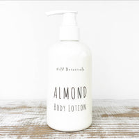 A white pump bottle labeled "Wild Botanicals Almond Lotion" stands on a rustic wooden surface with a neutral background. The label features simple, minimalist typography, highlighting the inclusion of organic jojoba oil.