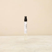 A small, transparent spray bottle with a black cap standing upright on a light beige surface, featuring a white label with minimalistic text and described as containing Norfolk Natural Living Parfum MidWinter 21 - 10ml.