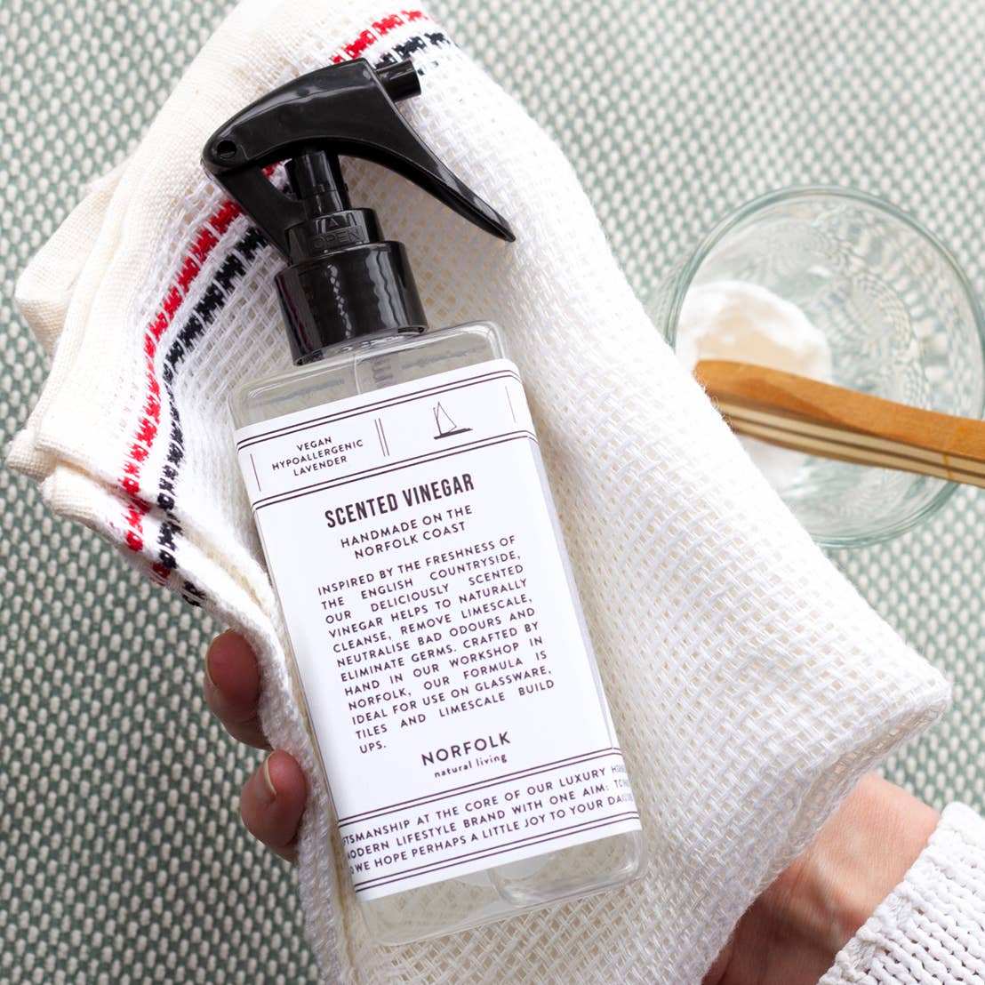 A hand holds a white textured cloth, a bottle of Norfolk Natural Living Rose Garden Scented Vinegar, and a small glass bowl with a wooden spoon and white powder, likely baking soda, on a light surface.