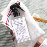 A hand holds a white textured cloth, a bottle of Norfolk Natural Living Rose Garden Scented Vinegar, and a small glass bowl with a wooden spoon and white powder, likely baking soda, on a light surface.