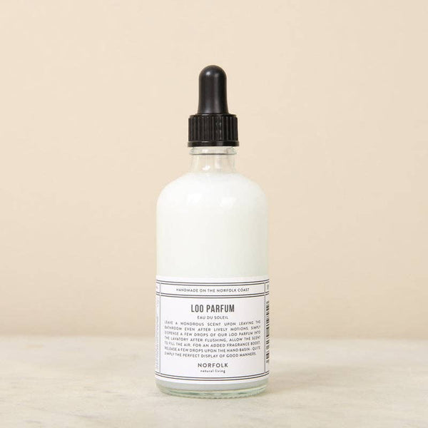 A clear glass dropper bottle with a black cap, filled with vegan white liquid labeled "Norfolk Natural Living Loo Parfum," rests on a light beige surface—a chic, natural fragrance solution for your bathroom.