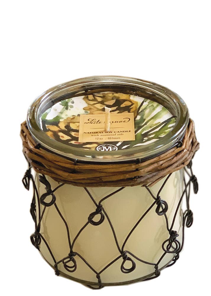 A Mangiacotti White Spruce Soy Farmhouse candle in a glass jar with a decorative wire and wicker casing. The jar has a metal lid featuring floral print beneath. A label on the lid reads "Lake Ground" and "Natural Soy Candle, 16oz." Infused with refreshing scents, this candle brings the essence of a woodland oasis into your home.