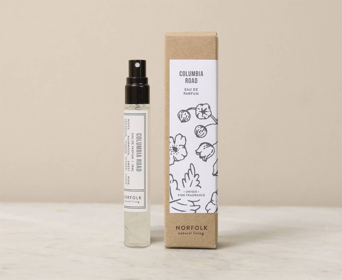 A 10ml glass spray bottle of Norfolk Natural Living Columbia Road Parfum, a vegan floral fragrance inspired by London's flower market, stands beside its beige and white box featuring floral line art and the brand’s logo.