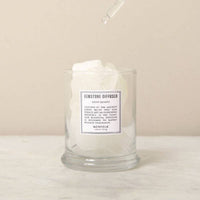 The Norfolk Natural Living Gemstone Diffuser Snow Quartz features a clear glass jar filled with large white aromatherapy stones, with a dropper above releasing liquid onto the crystals. The scene is set on a marble surface with a neutral background.