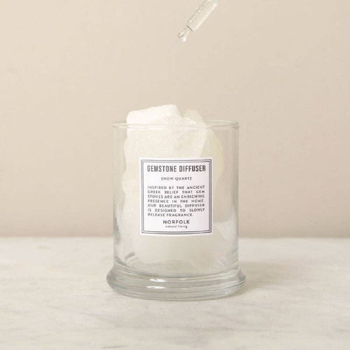 The Norfolk Natural Living Gemstone Diffuser Snow Quartz features a clear glass jar filled with large white aromatherapy stones, with a dropper above releasing liquid onto the crystals. The scene is set on a marble surface with a neutral background.