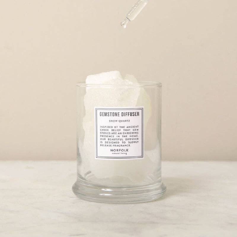 The Norfolk Natural Living Gemstone Diffuser Snow Quartz features a clear glass jar filled with large white aromatherapy stones, with a dropper above releasing liquid onto the crystals. The scene is set on a marble surface with a neutral background.