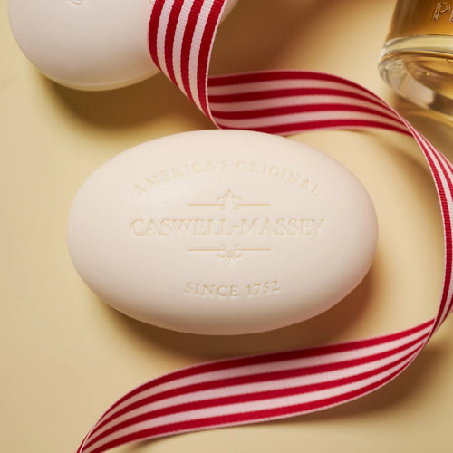 A Caswell-Massey Almond Three-Soap Set bar, embossed with the brand’s name, sits on a light surface beside a red-and-white striped ribbon and a partially visible bottle.