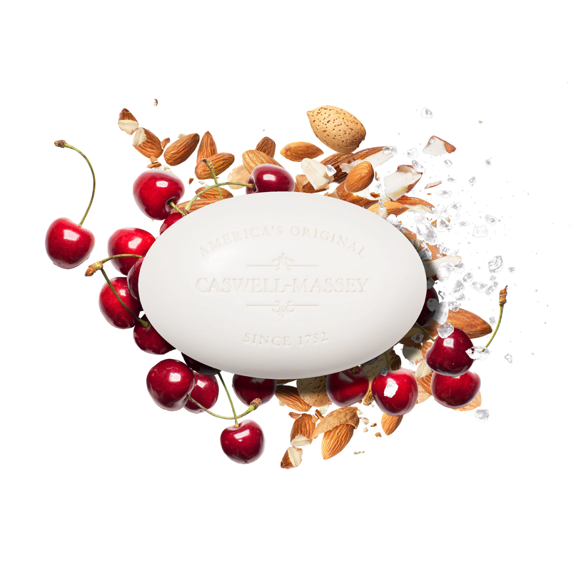 A white oval Caswell Massey Almond Bar Soap is surrounded by fresh cherries, almonds, almond shells, and sea salt on a white background, highlighting the moisturizing lather of this triple-milled soap.
