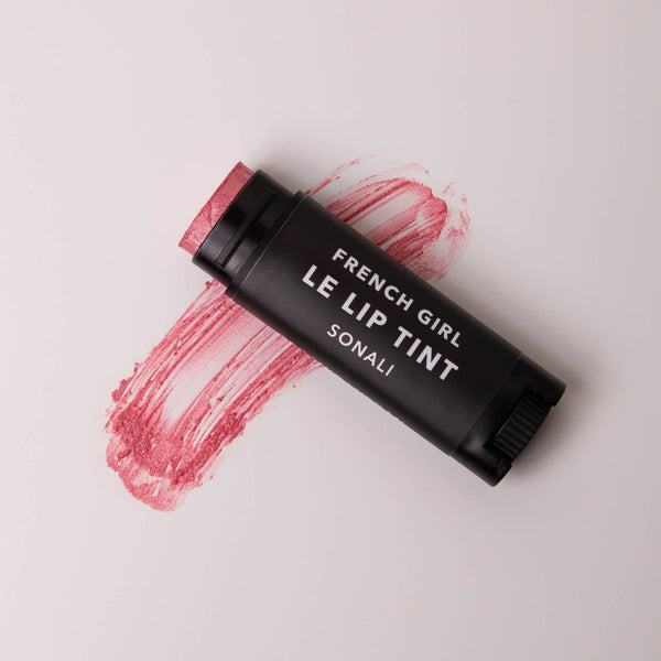 A black tube of FRENCH GIRL Tinted Lip Balm in the shade "Sonali" rests on a white surface, with a pinkish-red swatch of the tinted balm brushed beneath it.